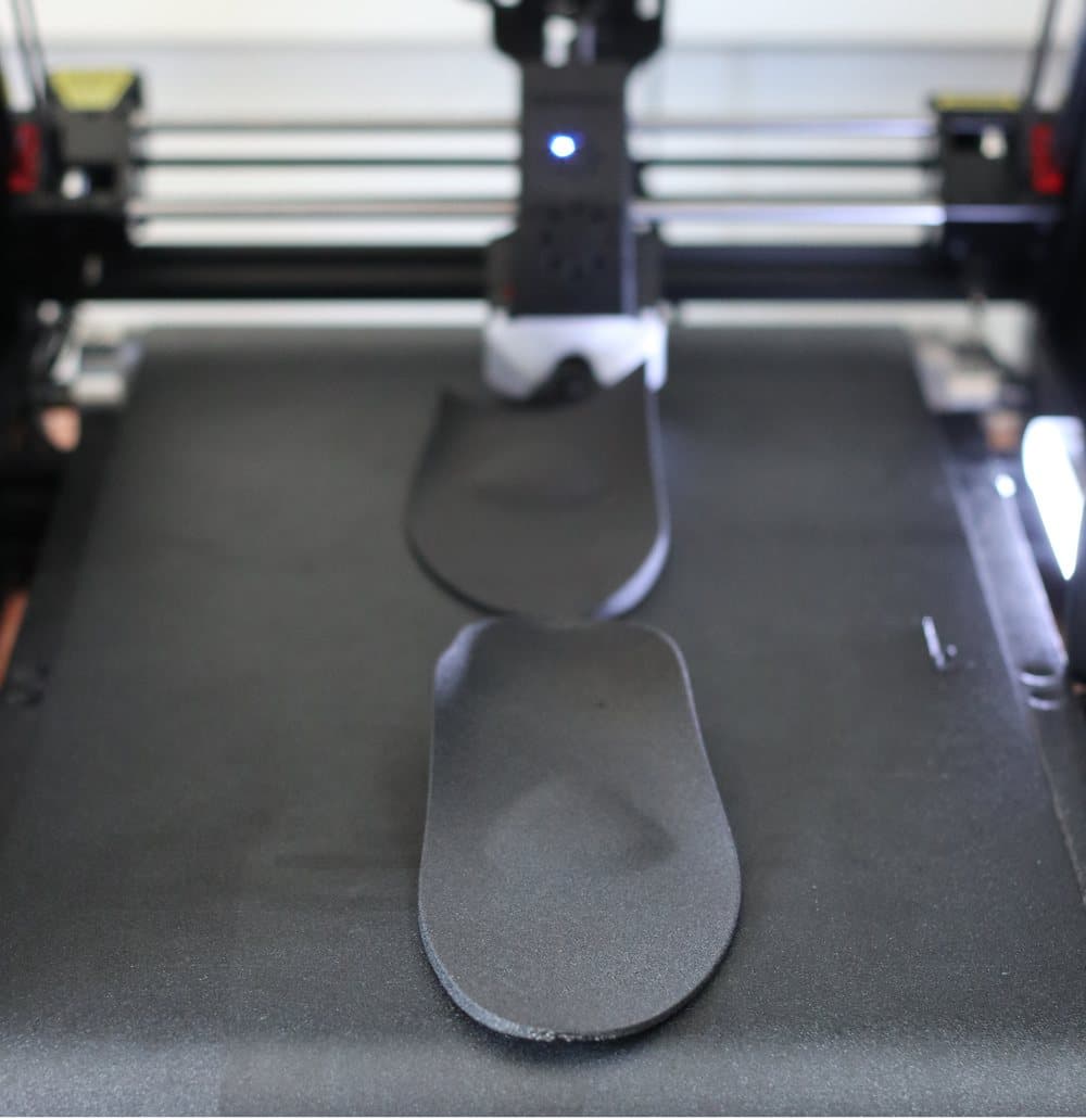 A 3D printer building an insole: the print head above the bed, with the shaped orthotic taking form beneath it.